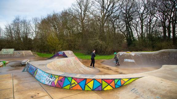 A concrete skatepark surrounded by trees, decorated with a colourful stained-glass effect mural.