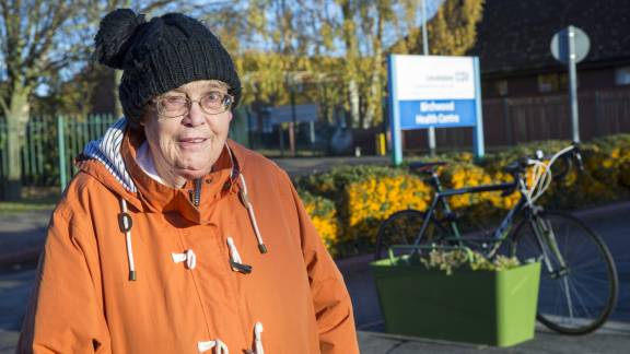 An elderly woman with glasses wears an orange raincoat coat and a black wool hat. Behind her is an open space with trees against a blue sky, a building, a wooden fence and a blue and white sign that reads "Birchwood Health Centre". There is also a topiary bush with yellow berries in it, and a blue bicycle attached to a green planter.
