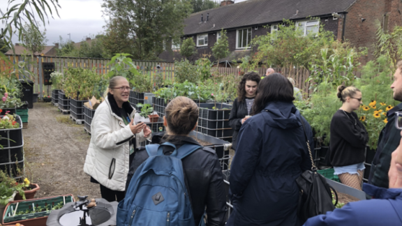 A group of volunteers, wearing waterproof jackets and coats, stand in a community garden. They are surrounded by multiple growing beds, filled with different plants. Most people are facing one woman who is speaking to the group.
