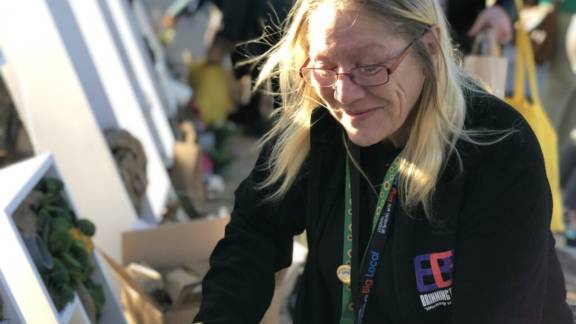 A woman with long blonde hair, glasses, wearing a black fleece and a green lanyard looks down at her hands out of frame.