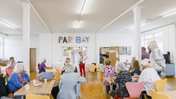 A group of people listening to a woman speak in a community hub.