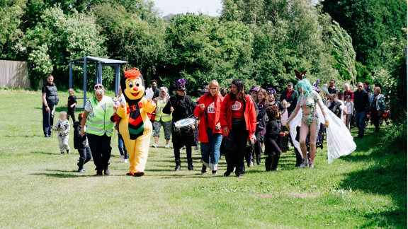 A green space with a crowd of people walking towards the camera, some in fancy dress.