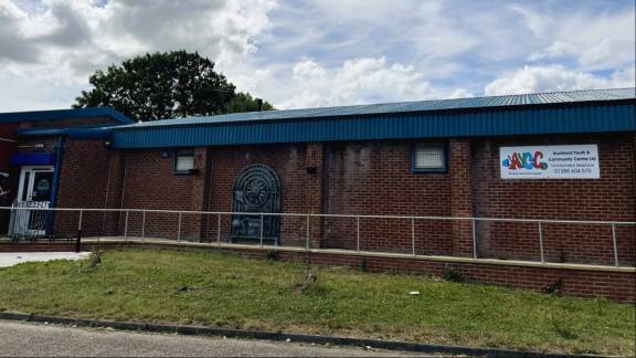 A single story red-brick building behind handrails with grass to the fore.