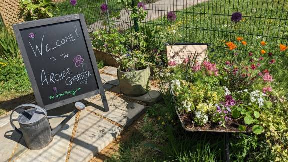 A floral display in a wheelbarrow in a garden, with a chalkboard sign reading 'Welcome to the Archer Garden.'