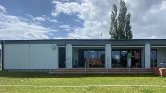 Sports pavilion on a sunny day, with two men on the steps chatting and holding coffee.
