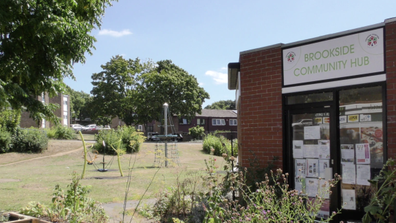 Brick building of Brookside Community hub with leaflets in the window and flowers and playground equipment outside.