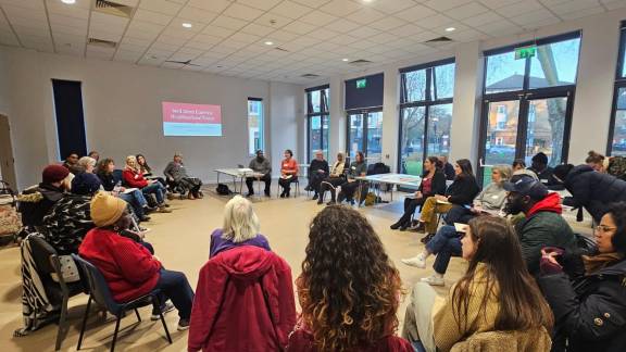 A large group of people sitting on chairs set out in a circle, in a bright room with large windows.