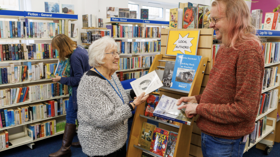 Interior of a library showing a woman and a man engaged in conversation near a wooden book display stand marked 'LOCAL AUTHORS' with a yellow sign. The woman on the left is elderly with white hair, glasses, and wearing a gray cardigan with a blue lanyard, holding a book. The man on the right has shoulder-length blonde hair, glasses, and is wearing a red knitted jumper, also holding a book. Behind them are extensive bookshelves with blue section markers reading 'Fiction - General' and 'Large Print - Fiction.' Another person can be seen browsing books in the background.