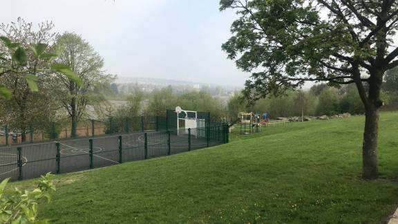 Landscape view of a mini multi-sports court and a playground, with grass and trees in the foreground.