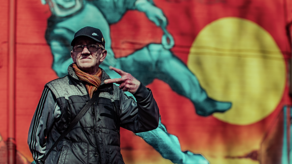 Man wearing glasses, a cap, and a jacket, standing in front of a colourful mural with a red background, yellow sun and blue spaceperson, making a peace sign.