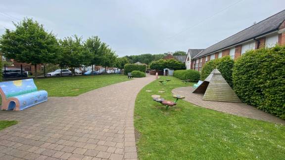 Brick pathway in a green space with a painted blue bench, toadstools and wooden teepee playhouse