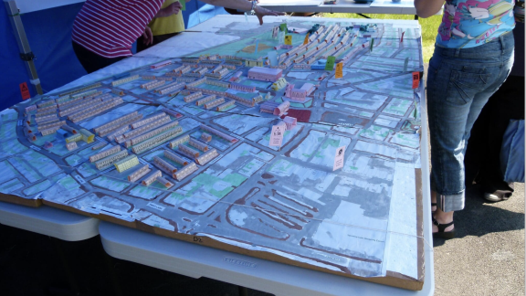 A giant 3D map of housing plans is set out across two trestle tables, surrounded by three people leaning over it. Their waist upwards is cropped. They are standing under a blue gazebo.