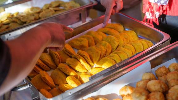 Steel warming dishes filled with pastries and fried food, with two people pointing at food items.