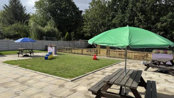 Wooden picnic tables with parasols in the sunshine in a paved area around a grassed play area.