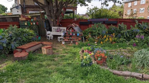 A woodland garden area under the shade of an old tree, with seating and flowers.