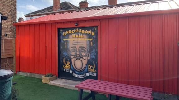 A red shed with 'Brocklebank Hall' painted on the doors along with an image of Ian Brocklebank.