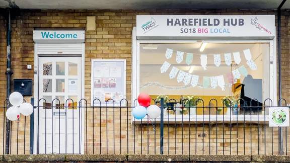 Yellow brick building with a white door and 'welcome' sign and a large window with bunting and a 'Harefield Hub: SO18 Big Local' sign.