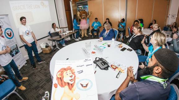 Two young present to a group of people in a room along with a person wearing a lion costume and a tutu.