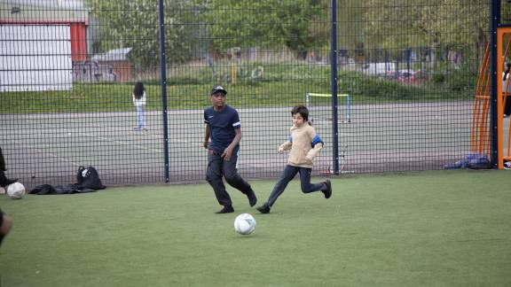 Two people kicking a football in a fenced-off sports area.