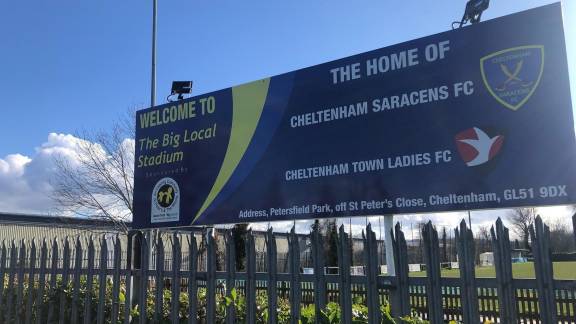 A large sign behind a fence reading 'The home of Cheltenham Saracens FC.'
