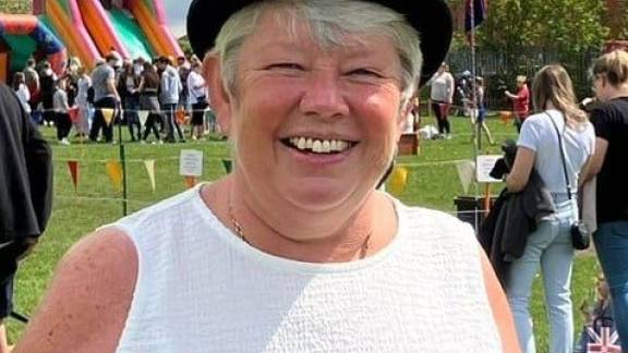 A woman wearing a black bowler hat, white tank top and a blue rosette, smiles at the camera. There is a bouncy castle in the background, bunting and crowds of people standing in a field.