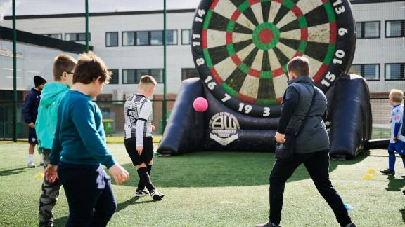 A group of children kicking a football towards a goal, which is a giant inflatable dart board.