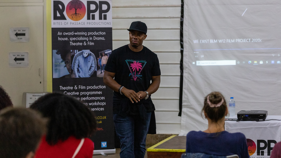 A man in a black t-shirt and cap standing beside a large banner for "ROPP - Rites of Passage Productions," describing it as an award-winning production house specializing in drama, theatre and film. Three people are sitting facing towards the man.
