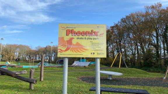 A sign reading 'Phoenix' in front of a play park and a bench.