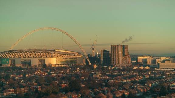 A landscape photo of Wembley at sunset. The Wembley Arena can be seen on the left of the horizon, with high rise buildings, cranes and lower buildings on the right. In the foreground are rows and rows of houses and trees.