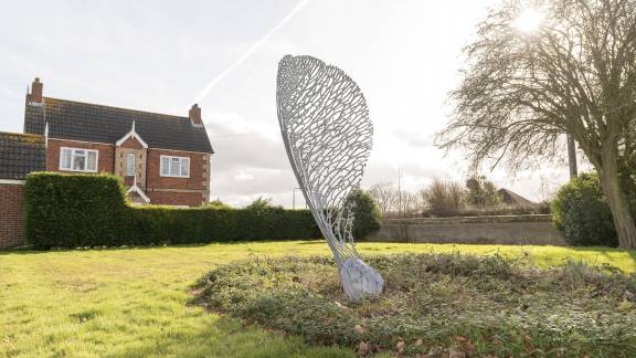 A large metalwork sculpture of a sycamore seed in a grassy area with a house in the background.