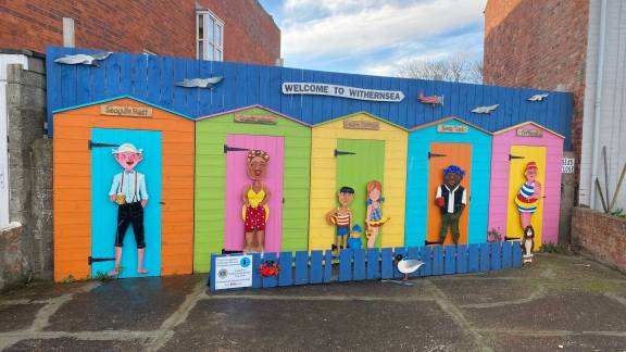 A sign reading "Welcome to Withernsea" sits on large wooden art wall, depicting seagulls flying above a set of five named beach huts with diverse characters standing in front of them.