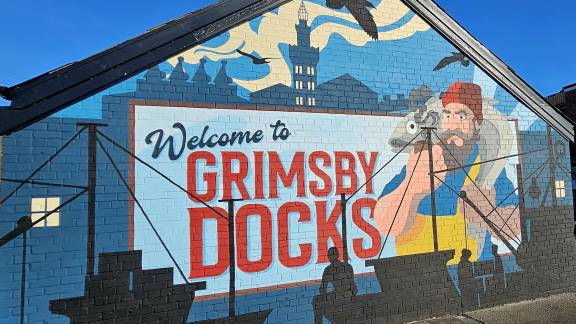 A painted mural on the side of a brick building depicts Grimsby Docks, with a painted sign across the middle that reads 'Welcome to Grimsby Docks' and a fisherman character with a fish around his neck.