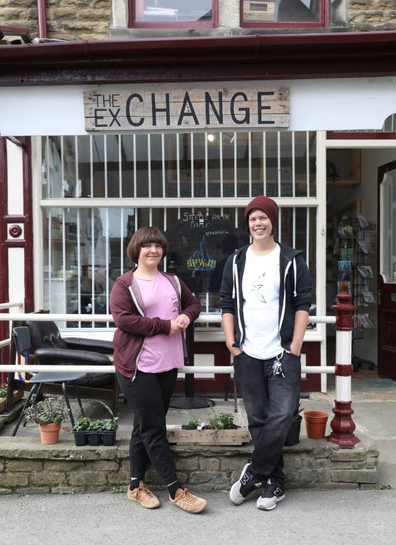 Two young people stand smiling in front of a traditional stone building with red-trimmed windows and white security gates. Above them hangs a wooden sign reading "THE EXCHANGE." The person on the left wears a pink t-shirt with a burgundy cardigan, while the person on the right wears a white t-shirt, dark hoodie, and burgundy beanie. Small potted plants and planters are arranged at the base of the building's stone foundation.
