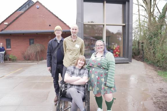 A group of people smiling at the camera, in front of a brick building on the left and a rectangular modern glass building on the right. One man with glasses wears a black coat, black trousers and a yellow beanie. Another man has grey facial hair and a beige shirt on. He stands behind a woman in a wheelchair who has mid-length blonde hair, grey tights and a tartan black and white dress. Her arms and legs are crossed. Next to her stands a woman with purple hair and glasses in a green dress with red and yellow mushrooms on it, wearing green knee-high socks, a green stripey cardigan.