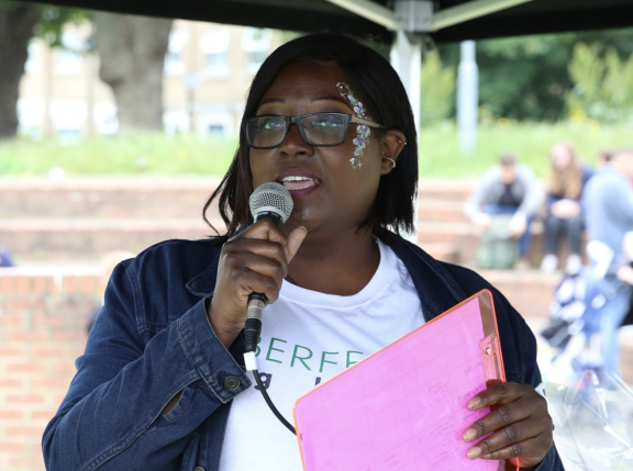 A woman wearing glasses and purple, white and green face paint on the side of her face speaks into a microphone. She is standing under a black gazebo, holding a pink folder. She wears a white t-shirt and a denim jacket. In the background, people sit on brick steps. A lawn, bushes, trees and a block of flats can be seen in the distance.