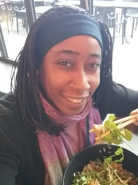 A woman with micro braids wearing a black hairband sits whilst eating food with chopsticks and smiles.