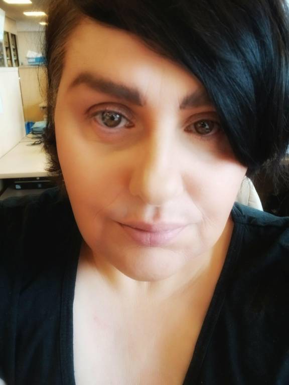 Close-up of a woman with dark hair swept to the side, wearing a black top, looking directly at the camera in a softly lit office environment.