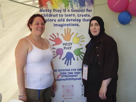 Two women standing together in front of a colourful promotional banner for 'Messy Play' sessions. The banner features rainbow-colored handprints arranged in a circle around the text 'MESSY PLAY' and explains that these are sessions for children to learn, create, explore and develop their imaginations. The woman on the left is wearing a white tank top and has reddish-brown hair, smiling at the camera. The woman on the right is wearing a black hijab and dark clothing with what appears to be an ID badge. Pink and blue balloons are visible in the background.
