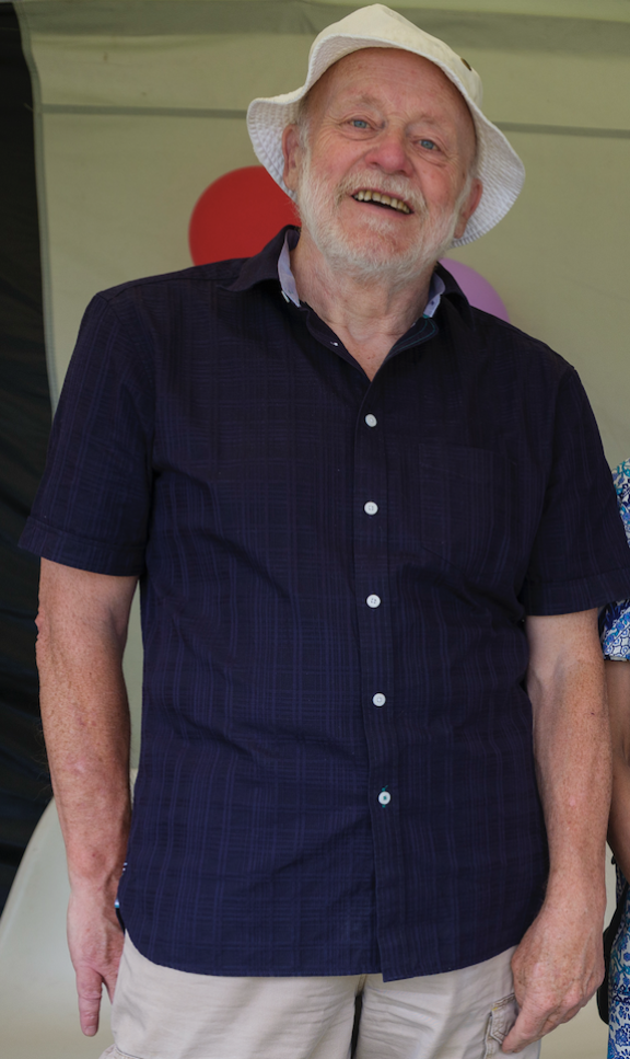 Man with a short white beard, wearing a navy, buttoned shirt and a white hat stands with his hands in his pockets and smiles.