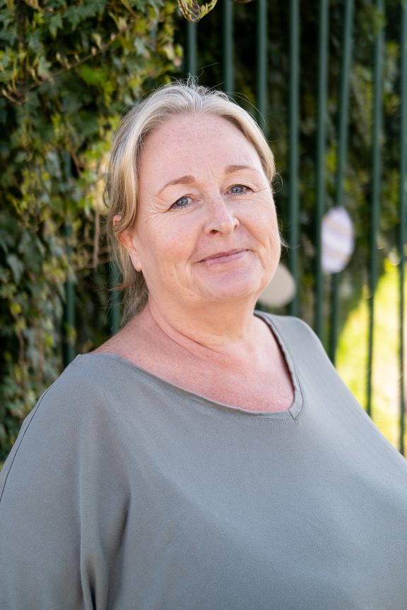 A shoulder-height portrait of a blonde woman with blue eyes, wearing a loose green t-shirt. She looks into the camera. Behind her is a green fence and ivy.