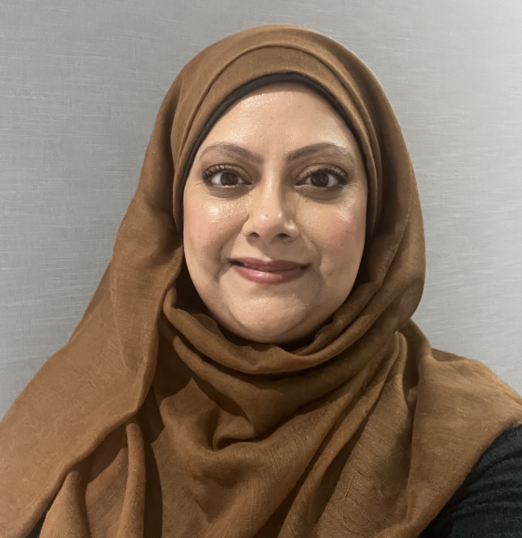A smiling woman stares straight at the camera. She is wearing a brown coloured hijab.