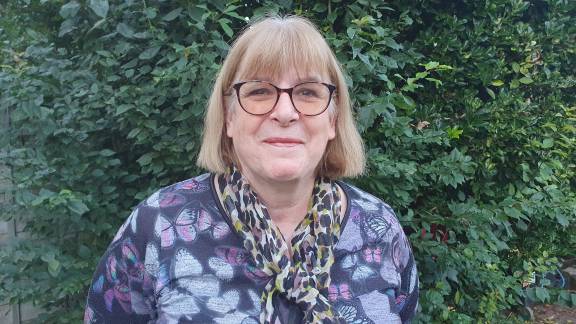 A middle-aged woman with shoulder-length blonde hair and a fringe stands smiling in front of green bushes. She wears dark-rimmed glasses and is dressed in a purple top with a colorful butterfly pattern . Around her neck is a leopard print scarf.
