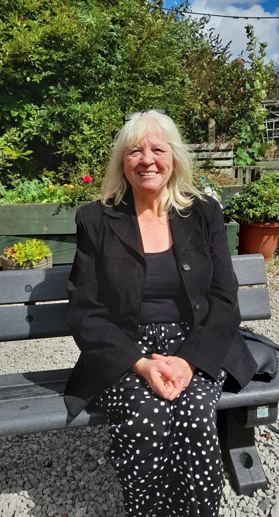 An older woman with shoulder-length blonde hair sits smiling on a dark wooden bench in a sunny garden setting. She wears a black blazer over a dark top, paired with a black skirt decorated with white polka dots. Her hands are folded in her lap as she poses for the camera. Behind her, lush green foliage and trees create a natural backdrop, with terracotta plant pots and garden features visible beneath a blue sky with white clouds.