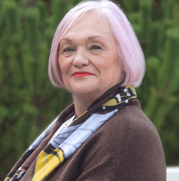 An older woman with a stylish short silver-pink bob haircut smiles warmly at the camera in a close-up portrait. She wears bright red lipstick and small earrings, and is dressed in a brown cardigan with a colorful scarf featuring yellow, blue, white, and brown patterns draped around her neck. The background shows soft-focused green foliage.