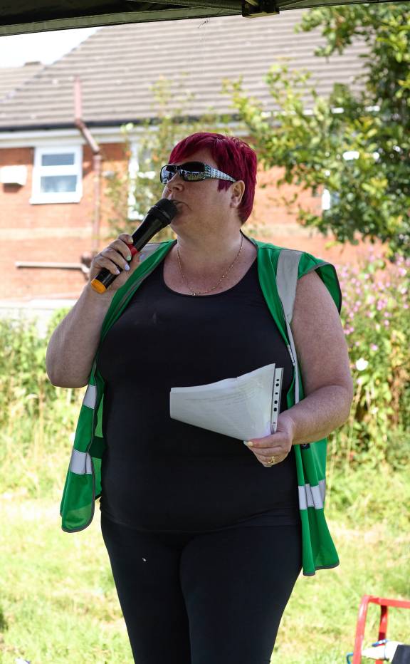 A woman wearing a black outfit and a green high-vis vest and sunglasses speaks into a microwave while holding a piece of paper. She has short cropped red hair. In the background, there is sunlit green grass and a brick residential house and a tree.