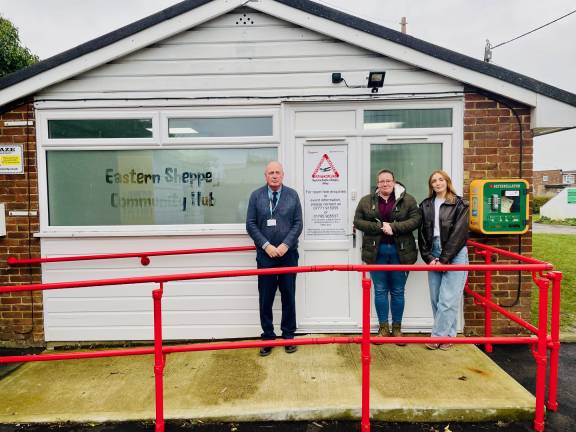 A man and two women stand outside a small brick and shiplap community building facing the camera. The window on the front of the building features frosted glass with the text 'Eastern Sheppey Community Hub' engraved on it. A red railing and accessible ramp lead up to the front door.