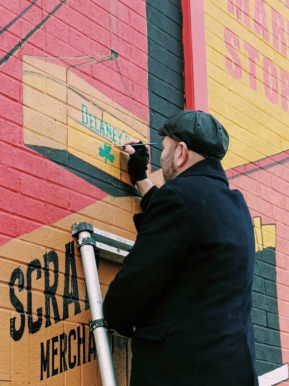 A man wearing a black coat and flat cap stands on a ladder painting details onto a colourful mural on a brick wall. The mural includes an illustration of a wooden crate labelled “Delaney’s” with a small green shamrock, along with large painted lettering on the wall.