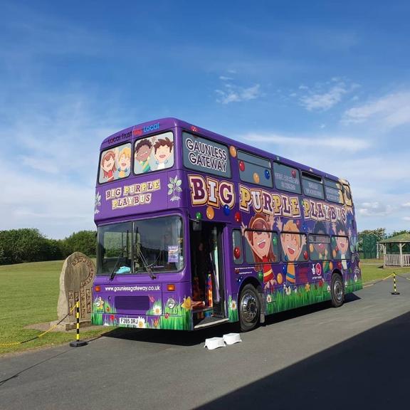 A large, bright purple double decker bus is parked next to a big green space. The text on the front of the bus reads 'Local Trust Big Local' and down the side reads 'Big Purple Playbus' and 'Gaunless Gateway' in playful font. There are illustrations of children cheering painted onto the bus.
