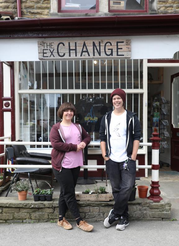 Two young people stand smiling in front of a traditional stone building with red-trimmed windows and white security gates. Above them hangs a wooden sign reading "THE EXCHANGE." The person on the left wears a pink t-shirt with a burgundy cardigan, while the person on the right wears a white t-shirt, dark hoodie, and burgundy beanie. Small potted plants and planters are arranged at the base of the building's stone foundation.