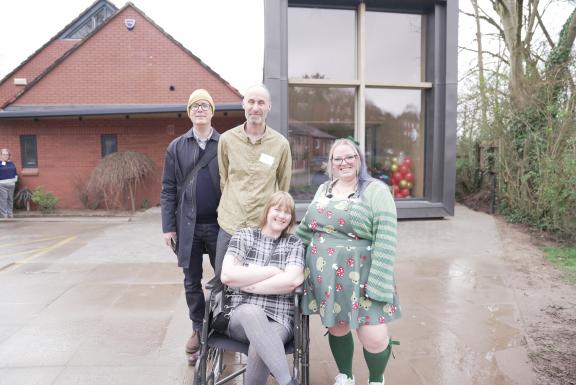 A group of people smiling at the camera, in front of a brick building on the left and a rectangular modern glass building on the right. One man with glasses wears a black coat, black trousers and a yellow beanie. Another man has grey facial hair and a beige shirt on. He stands behind a woman in a wheelchair who has mid-length blonde hair, grey tights and a tartan black and white dress. Her arms and legs are crossed. Next to her stands a woman with purple hair and glasses in a green dress with red and yellow mushrooms on it, wearing green knee-high socks, a green stripey cardigan.